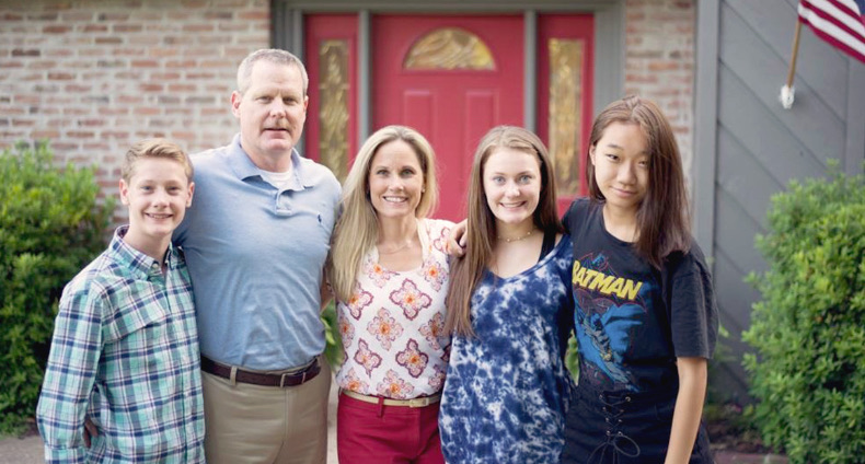 A host family of five standing together and smiling in front of a red front door.