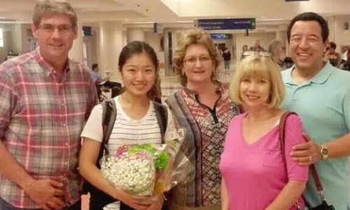 A host family welcoming a student holding a bouquet of flowers at an airport arrivals hall.