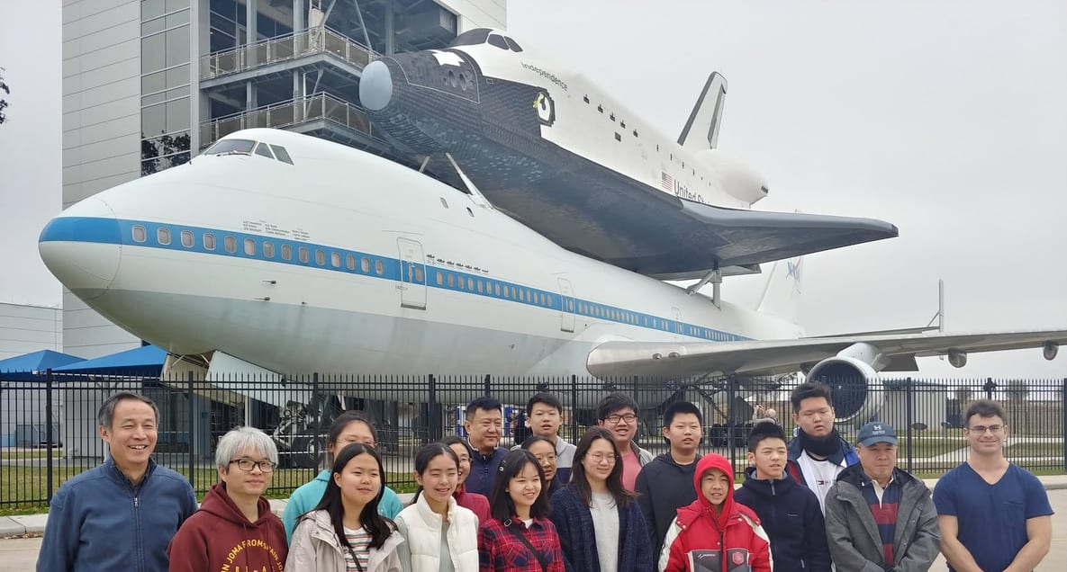 A group of students outdoors in front of a Space Shuttle carrier aircraft on display.