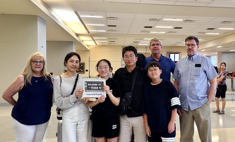 A group of students and adults standing together indoors holding a welcome sign.